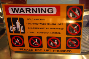 Photo of a sign on an escalator with visual and written instructions about how not to stand and what not to take on, including wheelchairs and prams.